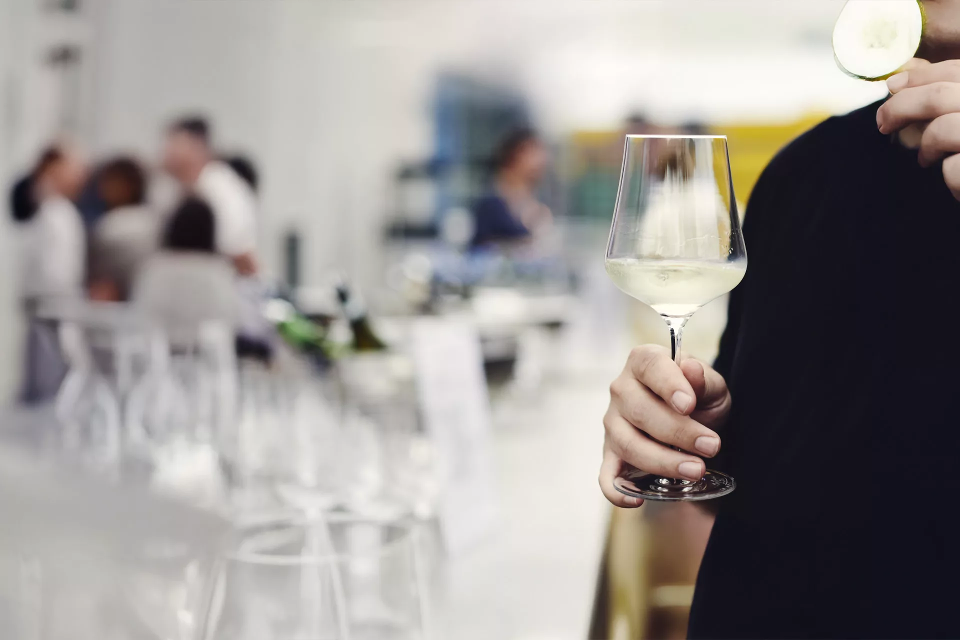 Person hält ein Glas Weißwein in einer Bar, im Hintergrund Gäste.